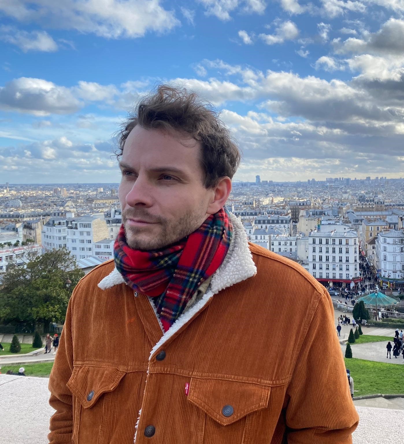 A man in a yellow jacket and tartan scarf stands in front of a city landscape.